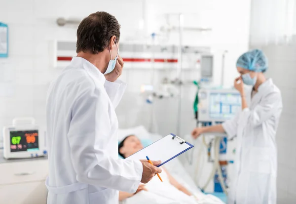 Médico com forma médica na mão olhando para a enfermeira de pé perto do paciente — Fotografia de Stock