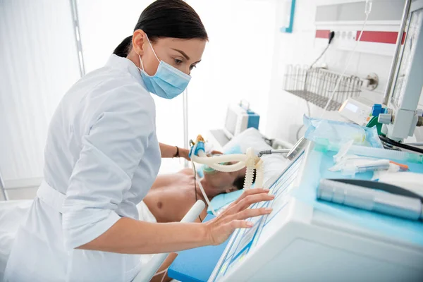 Médico joven revisando la máquina respiratoria mientras pone máscara de oxígeno en el paciente — Foto de Stock