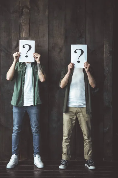 Young gay couple covering faces with question signs