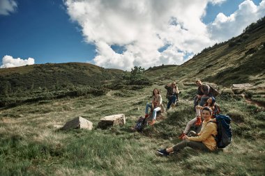 Mutlu beş turist turistik arka çantalar top birlikte takmak için yürüyüş ile. Dört kadın ve adam yere oturuyor ve tırmanma devam etmeden önce dinlenme