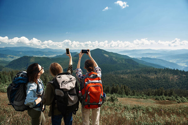 Female friends standing with focus on back and taking picture of amazing peaks scenery in distance while using smartphones