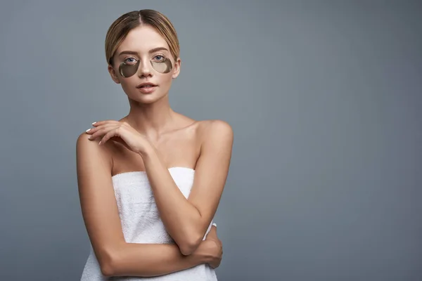 Calm woman wearing white towel and having eye patches on the face