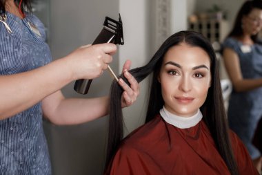 Sandalyede oturan ve iyi vakit güzellik Studio süre koruyucu elbise giyen gülümseyen kadın portresi. Kadın saç sanatçı yanında duran ve istemci penye