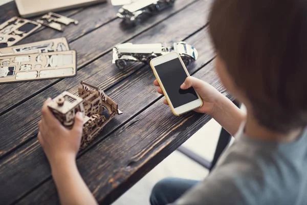 Rapaz progressivo sentado à mesa com construtor de madeira e segurando smartphone moderno em suas mãos — Fotografia de Stock