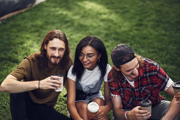 Trois jeunes amis dégustant un café assis sur l'herbe ensemble — Photo