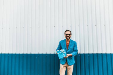 Güzel mevcut. Duvara yakın duran ve güzel mavi mevcut gösterilen süre gülümseyen şık giysiler içinde neşeli sakallı genç adam