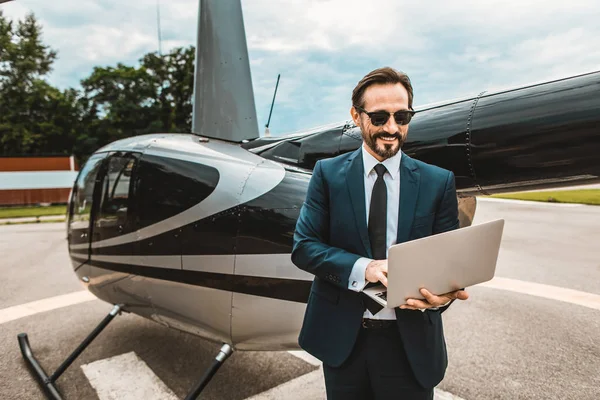Счастливый бизнесмен прячет современный ноутбук, стоя рядом с вертолетом — стоковое фото