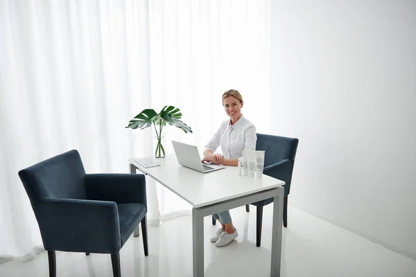 Hermoso cosmetólogo sentado a la mesa con portátil — Foto de Stock