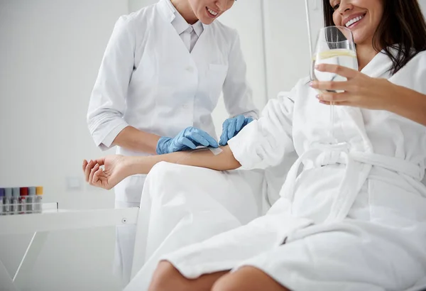 Medico controllando flebo endovenoso sulla mano della signora mentre beve acqua — Foto Stock