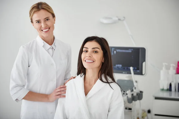 Cosmetologista e mulher morena posando no salão de beleza — Fotografia de Stock