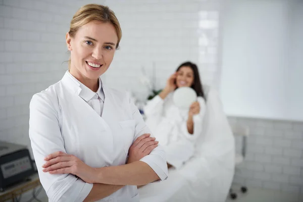 Cosmetologista alegre em pé em seu gabinete wile cliente descansando no leito do dia — Fotografia de Stock