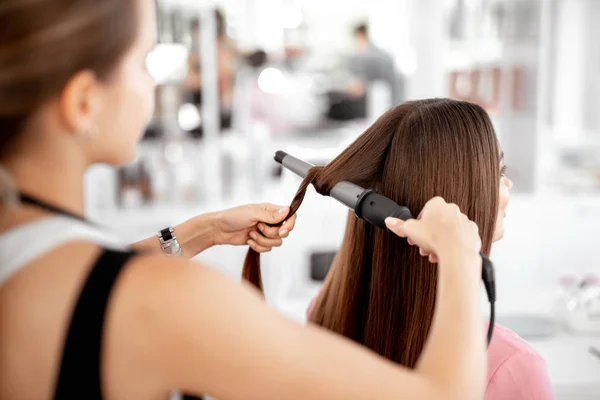 Jovem senhora sentado e cabeleireiro fazendo seu cabelo encaracolado — Fotografia de Stock