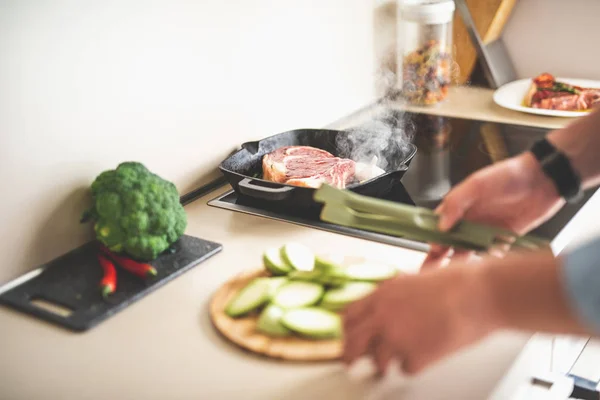 Piece of meat with steam frying on stovetop while man preparing vegetables
