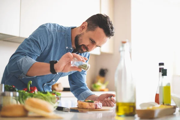 Jongeman specerijen toe te voegen aan vlees terwijl je in de buurt van keukentafel — Stockfoto