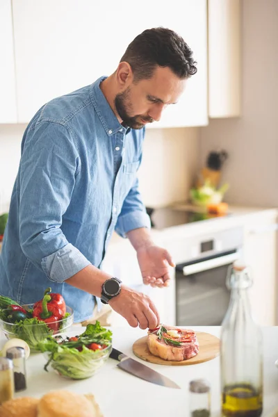 Jongeman kruiden toe te voegen aan vlees terwijl je in de buurt van keukentafel — Stockfoto