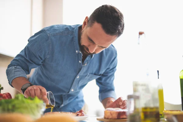 若い男の台所のテーブルの近くに立っている間肉を料理に焦点を当ててください。 — ストック写真