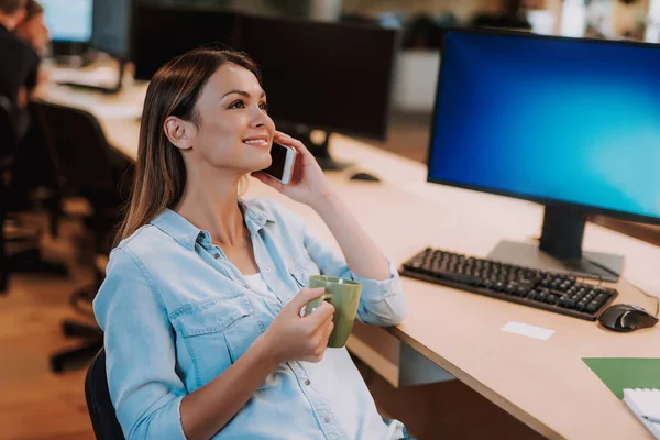 Mooie jonge dame met kop warme drank praten op mobiele telefoon op het werk — Stockfoto