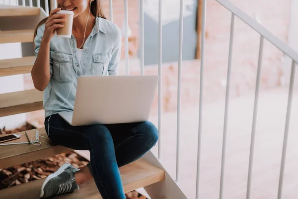 Charmig ung dam i hörlurar njuter av varm dryck medan du arbetar på laptop — Stockfoto