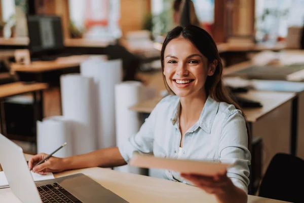 Fröhliche junge Frau, die im Büro mit Dokumenten und Laptop arbeitet — Stockfoto