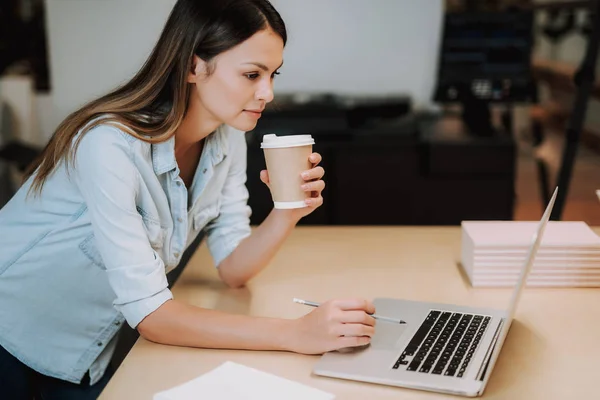 Schöne junge Frau mit Heißgetränk schaut auf Laptop — Stockfoto