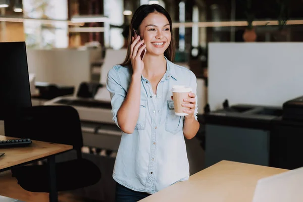 Vrolijke jongedame met warme drank met telefoongesprek op kantoor — Stockfoto