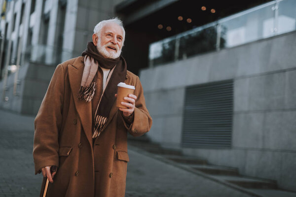 Stylish old man with hot drink walking on the street