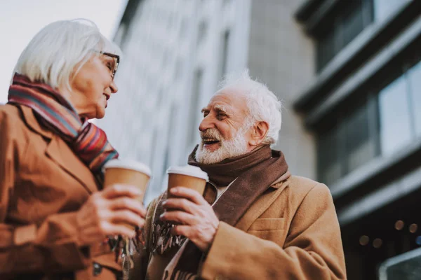 Side view portrait of stylish old gentleman in coat chatting with wife. They holding cups of coffee while looking at each other and smiling