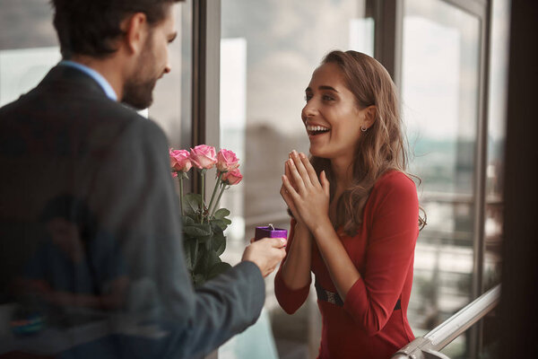 Concept of surprising engagement. Waist up portrait of young male holding box with ring and flowers while offering to marry him to his happy lover