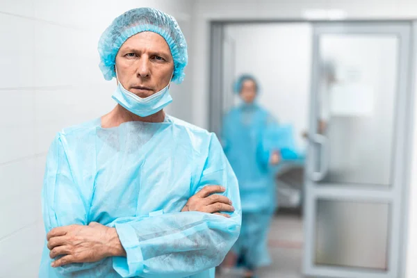 青い医療制服顔をしかめ一方腕を病院の廊下で立って越えで穏やかな専門の医者 彼の同僚のバック グラウンドの上を歩く — ストック写真