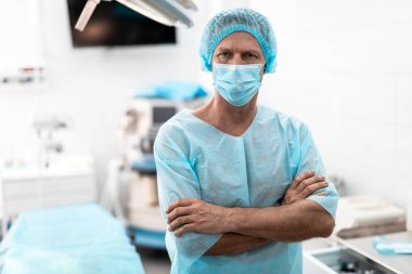 Ameliyat odasında duran çapraz kolları olan orta yaşlı cerrah