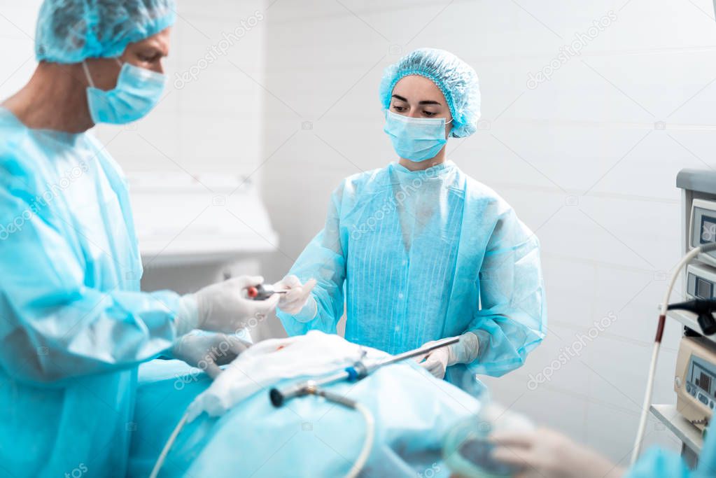 Young nurse in protective mask giving surgical instrument to surgeon