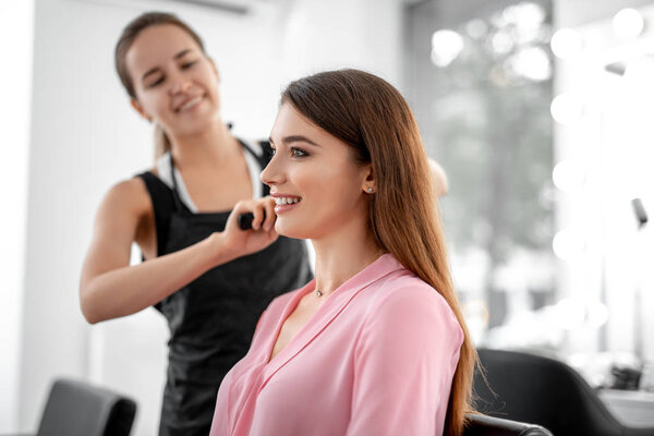 Waist up photo of beautiful smiling woman is sitting in beauty salon while female stylist making hair styling