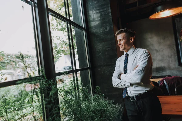 Concept of success and well-being. Waist up portrait of handsome smiling businessman staying in cafe and looking on window. Copy space on left