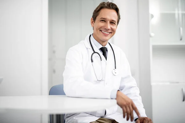 Derék fel boldog orvos ül az egyik kezével az asztalra — Stock Fotó