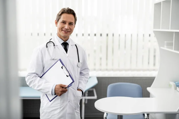 Médico general confiado poniendo una mano en el bolsillo y sonriendo — Foto de Stock