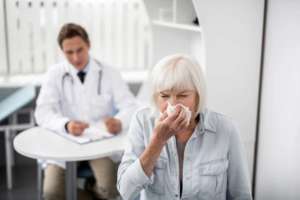 Mujer envejecida estornudando y médico general mirándola — Foto de Stock