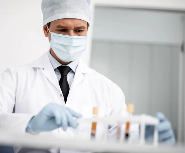 Médico sério de uniforme médico sentado e a olhar para os tubos de ensaio — Fotografia de Stock