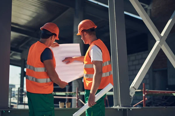 Constructor atento apuntando al dibujo mientras está de pie con su compañero de trabajo — Foto de Stock