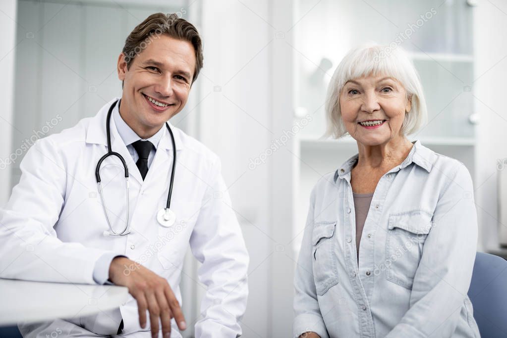 Positive young doctor looking glad while sitting next to the emotional elderly lady and smiling