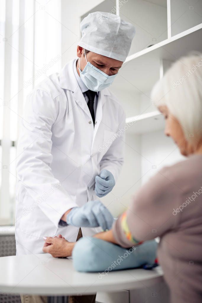 Careful professional doctor in medical mask standing in front of his patient and making a necessary injection