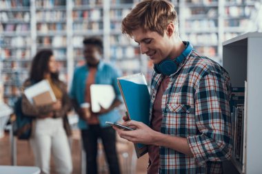 Genç adam onun smartphone kütüphanede ekran bakarak gülen