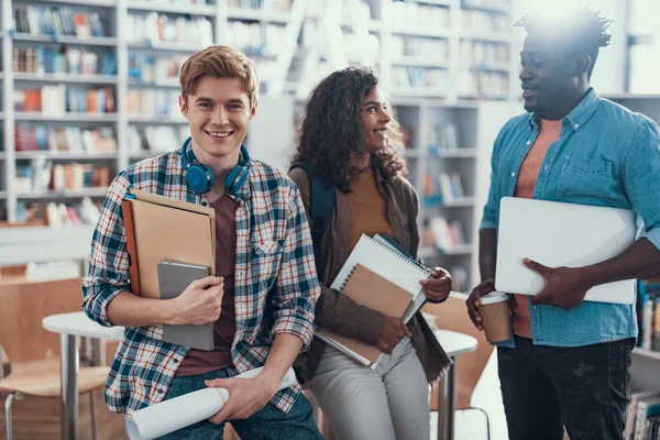 Três estudantes alegres sentados na biblioteca e sorrindo — Fotografia de Stock