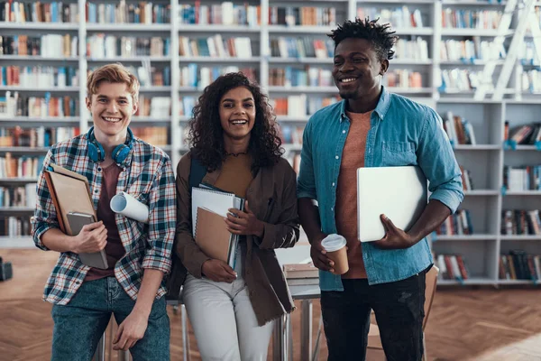 Drei glückliche Studenten, die in der Bibliothek sitzen und lächeln — Stockfoto