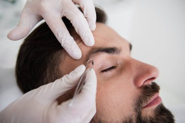 Hands in rubber gloves holding pair of tweezers and plucking eyebrows