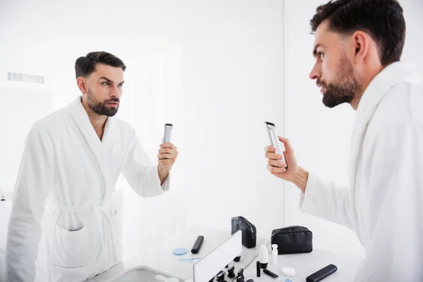 Cintura para cima de homem sério de pé com sua máquina de barbear no banheiro — Fotografia de Stock