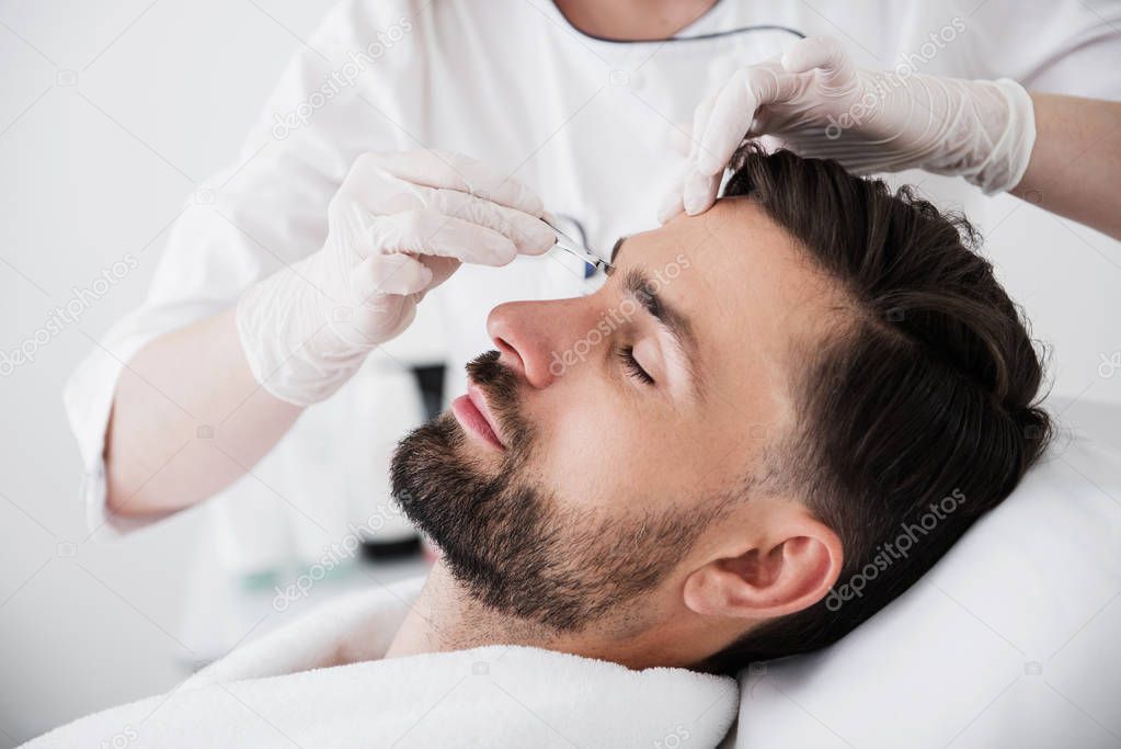 Bearded man sitting with closed eyes and experienced cosmetologist in white uniform and white rubber gloves plucking his eyebrows with tweezers