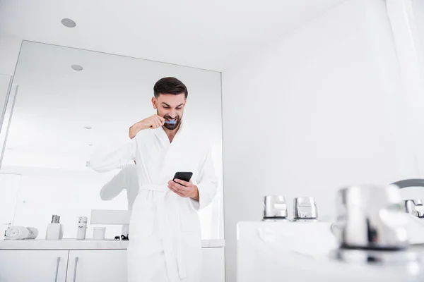 Взволнованный мужчина смотрит на свой смартфон и чистит зубы — стоковое фото