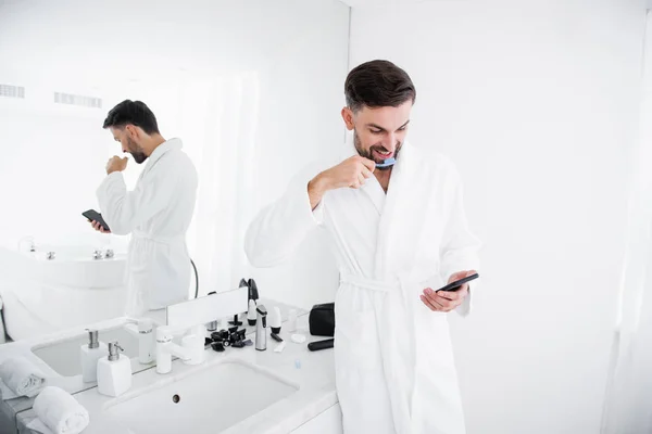 Hombre emocional cepillándose los dientes y mirando el teléfono inteligente — Foto de Stock