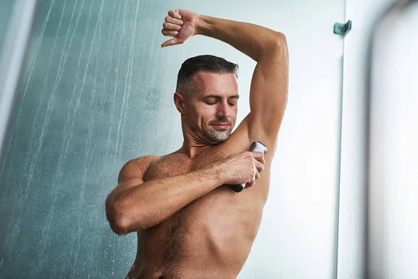 Мускулистый молодой человек бреет подмышки, принимая душ — стоковое фото
