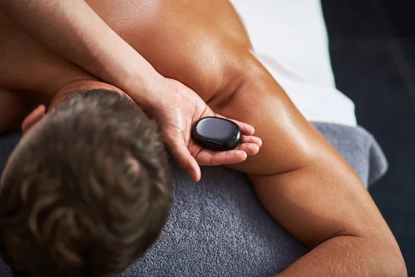 Молодий чоловік лежить на масажному столі, маючи масаж спини гарячим каменем — стокове фото